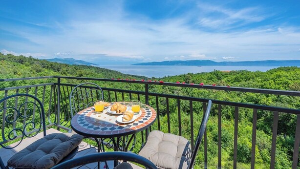 A balcony with a table set for breakfast, overlooking a lush green landscape and a distant body of water.