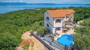 A two-story house with a swimming pool, surrounded by lush greenery and a view of the sea.