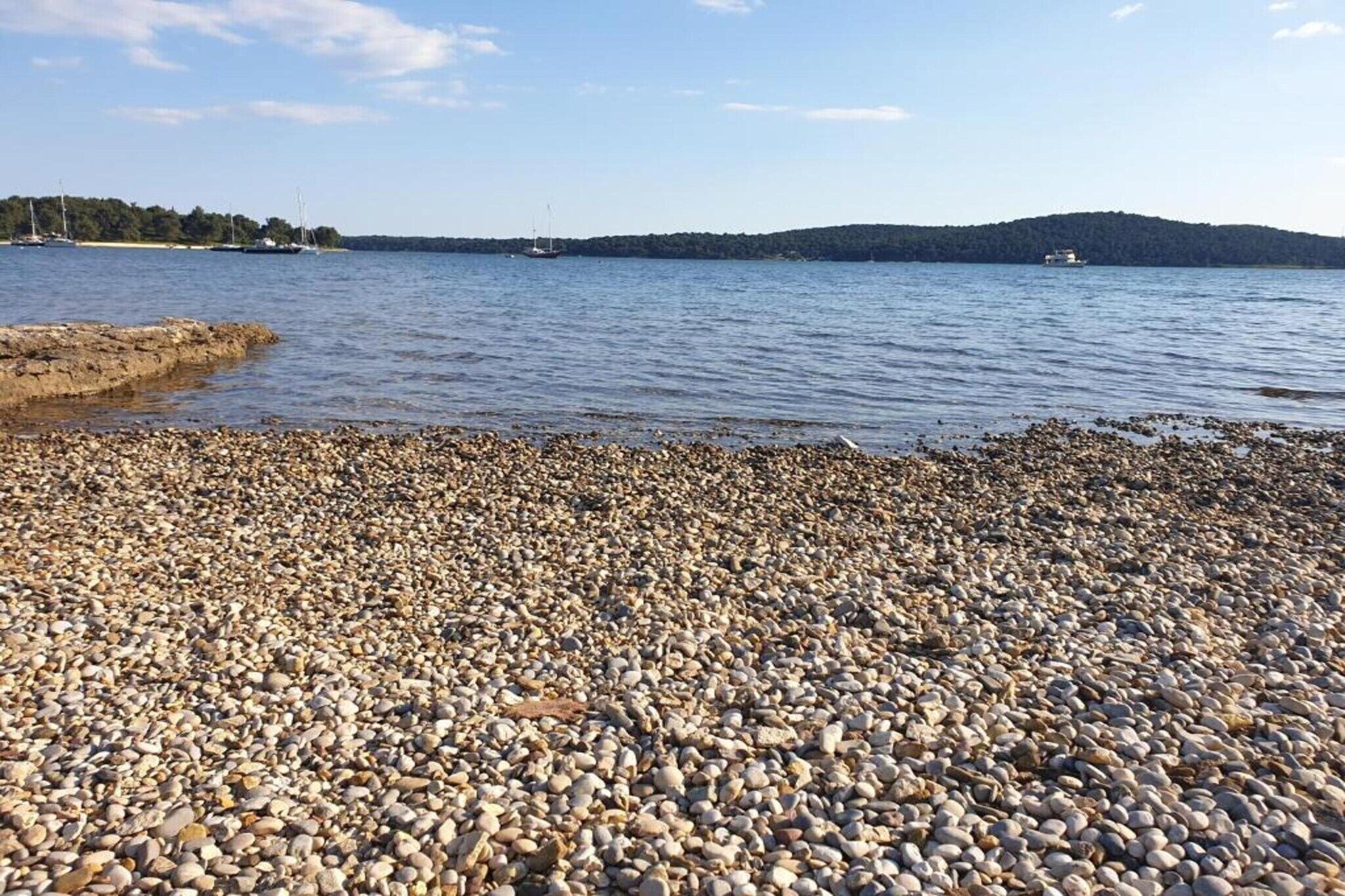 A pebbled beach with boats in the background and a forested hill.