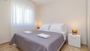 Una cama bien hecha con colcha gris, almohadas blancas y dos toallas blancas dobladas. Hay una mesita de noche blanca con una lámpara encima. La habitación tiene paredes blancas y una ventana con cortinas de gasa.