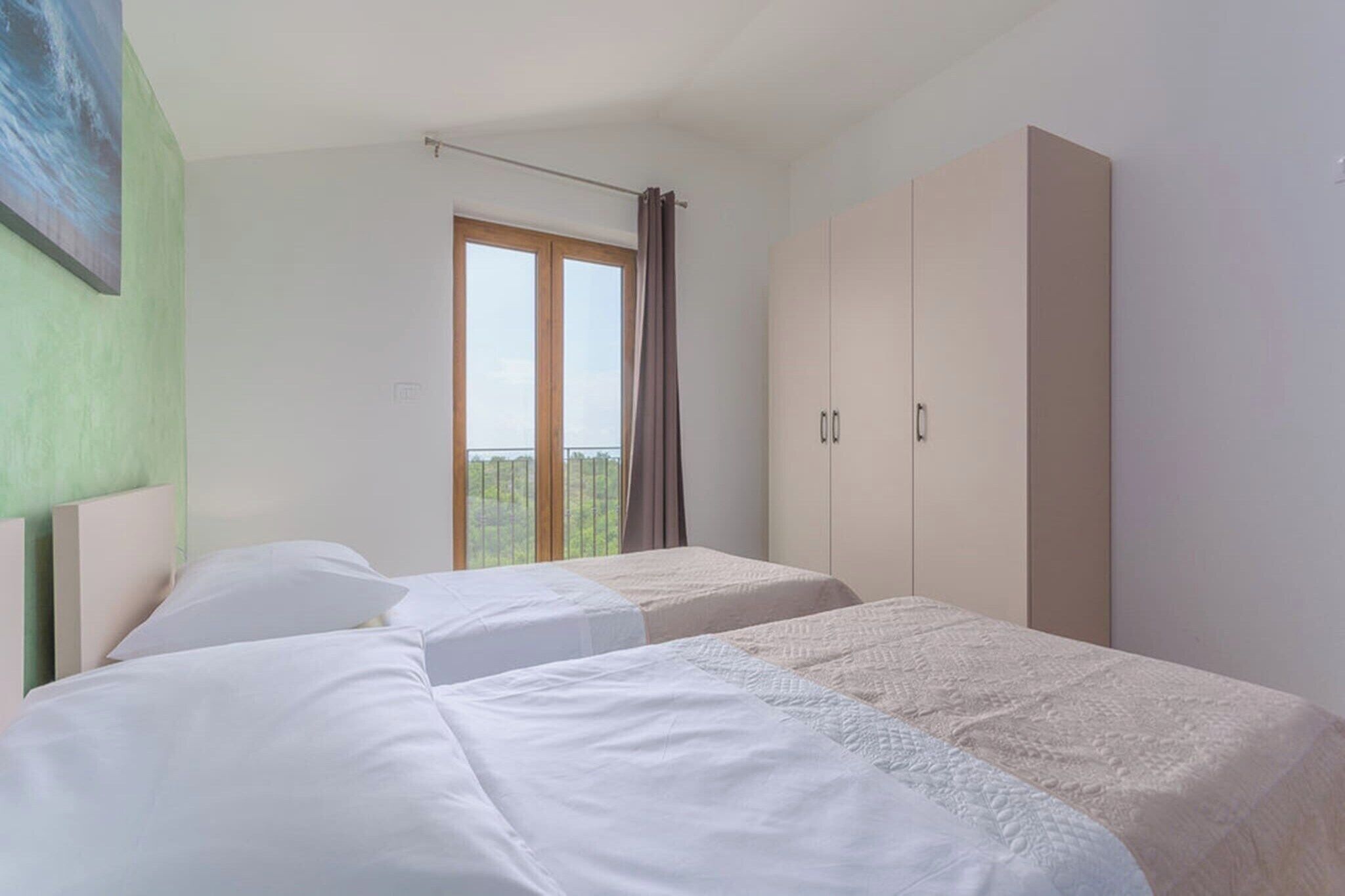 A bedroom with two beds, a large wardrobe, and a window with a view of greenery.