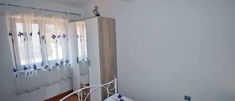 A bedroom with a bed, a window with sheer curtains, a wooden wardrobe, and a white metal bed frame.