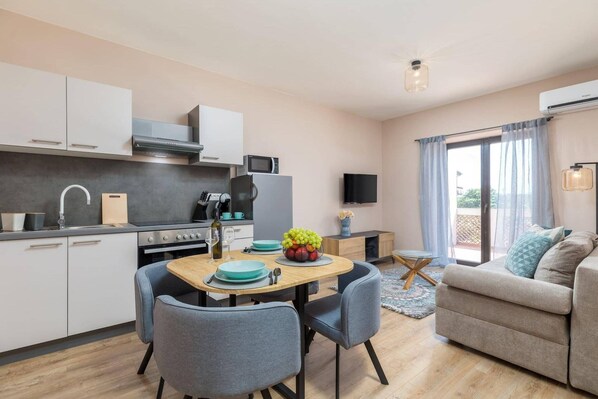A modern kitchen with white cabinets, a dining table with blue chairs, and a living area with a sofa and TV.