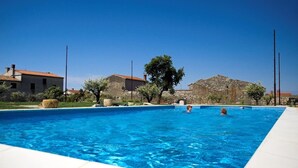A rectangular swimming pool with clear blue water, surrounded by stone buildings and a tree.