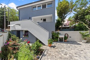 Een modern, tweelaags gebouw met een grijze gevel, een klein balkon en een betegeld terras met plantenpotten.