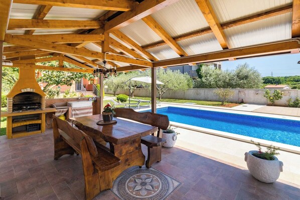 An outdoor patio with a wooden table and benches, a barbecue grill, and a pool.