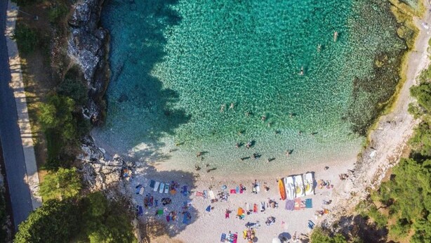 Vista aérea de una playa con agua turquesa cristalina, una orilla arenosa y varios sombrillas y sillas de playa coloridas.