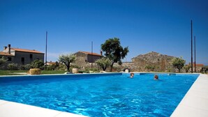 Una piscina rectangular con agua azul cristalina, rodeada de edificios de piedra y un gran árbol.