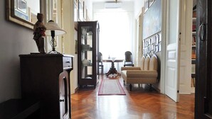 A hallway with wooden flooring, a lamp, a dresser, a mirror, and a rug leading to a room with a chair and a table.