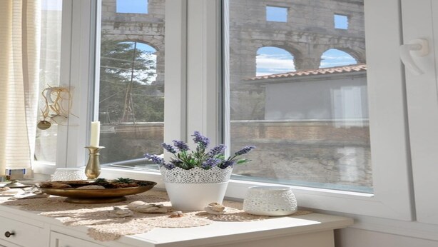 A window with a view of an ancient stone structure, a white vase with purple flowers, a candle, and a bowl on a window sill.
