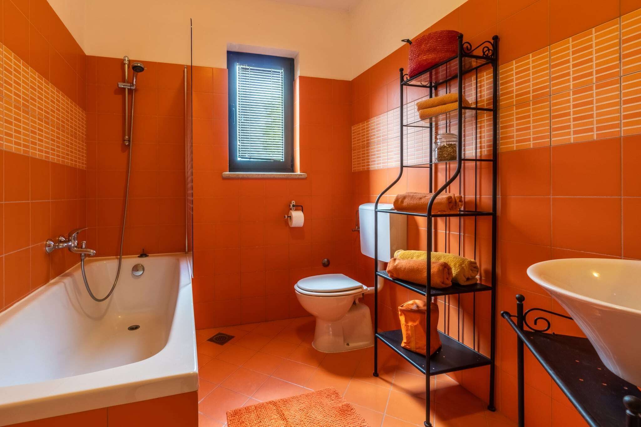A bathroom with an orange tiled wall, a white bathtub, a toilet, a shelf with towels, and a large white sink.