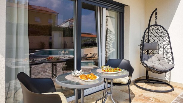 A balcony with a glass door, two round tables with plates of food, a hanging chair, and a view of a swimming pool and buildings. - Luxe villa in Pula with pool and jacuzzi (Pula)