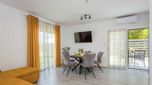 A modern living room with a sofa, a dining table with chairs, a flat-screen TV, and a sliding glass door leading to an outdoor area.