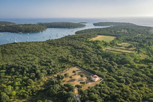 An aerial view of a coastal area with dense greenery, a small white building, and a marina with boats.