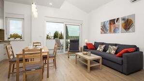 Un salon moderne avec un canapé gris, une table à manger en bois et des chaises. Trois œuvres d’art ornent le mur. Une porte-fenêtre coulissante donne sur l’extérieur, et on aperçoit un coin cuisine à travers la porte.
