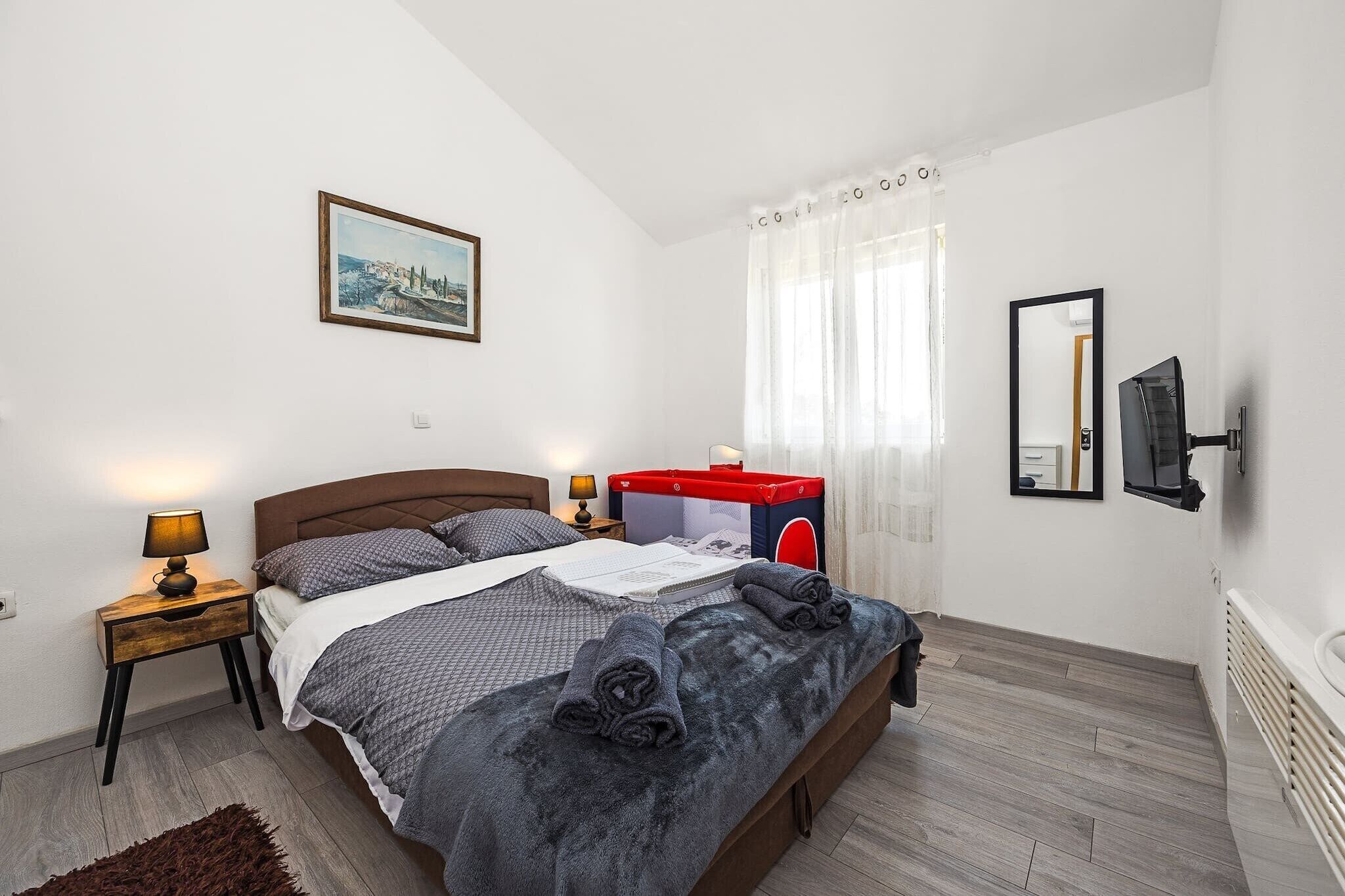 Une chambre à coucher comprenant un lit, des tables de chevet, une commode, un meuble rouge et bleu, un tableau, un miroir et une fenêtre avec des rideaux.