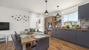 A modern kitchen with a dining area, featuring a wooden table, grey chairs, and a TV on the wall. - Modern Rscape with Pool (Baderna)