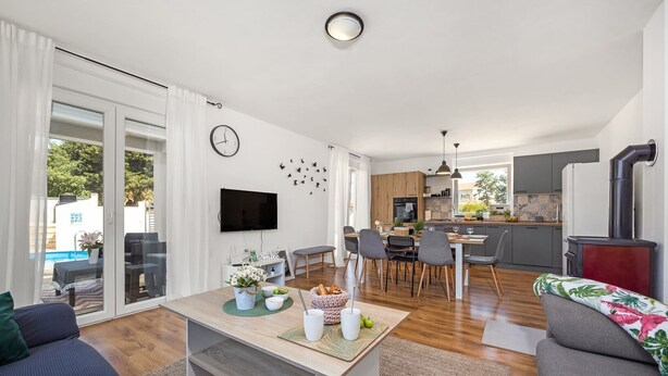 A modern living room with a sofa, a coffee table, and a TV. There is a dining area with chairs and a table. The kitchen is visible with cabinets and a stove. - Modern Rscape with Pool (Baderna)