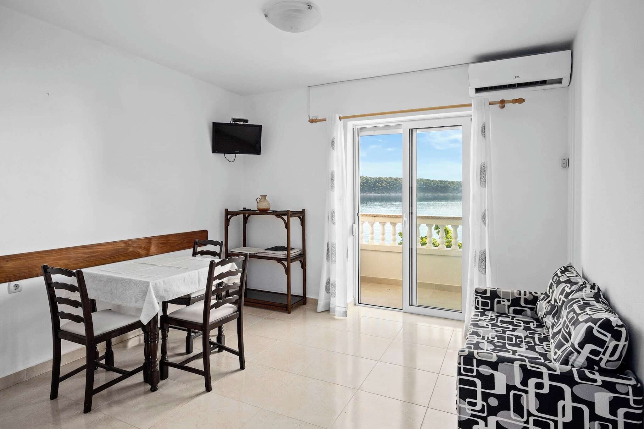A room with a dining table and chairs, a sofa, a TV mounted on the wall, and a sliding glass door leading to a balcony with a view of the sea.