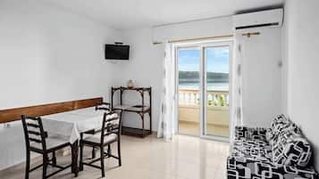 A room with a dining table and chairs, a sofa, a TV mounted on the wall, and a sliding glass door leading to a balcony with a view of the sea.