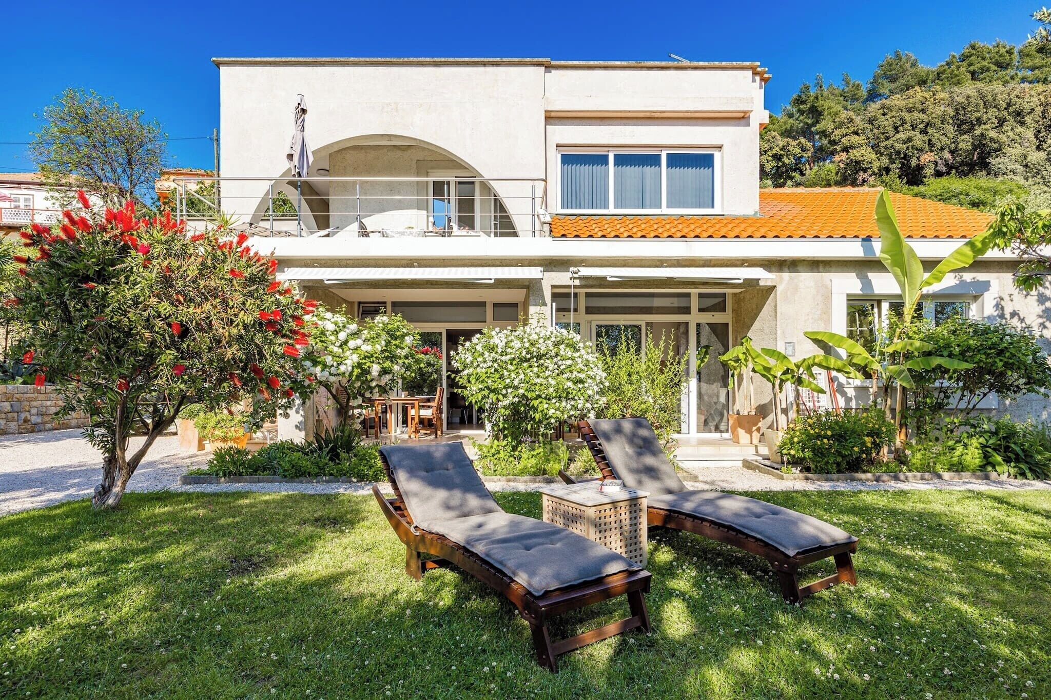 Une maison à deux étages avec un balcon, un jardin doté de chaises longues et un arbre aux fleurs rouges.