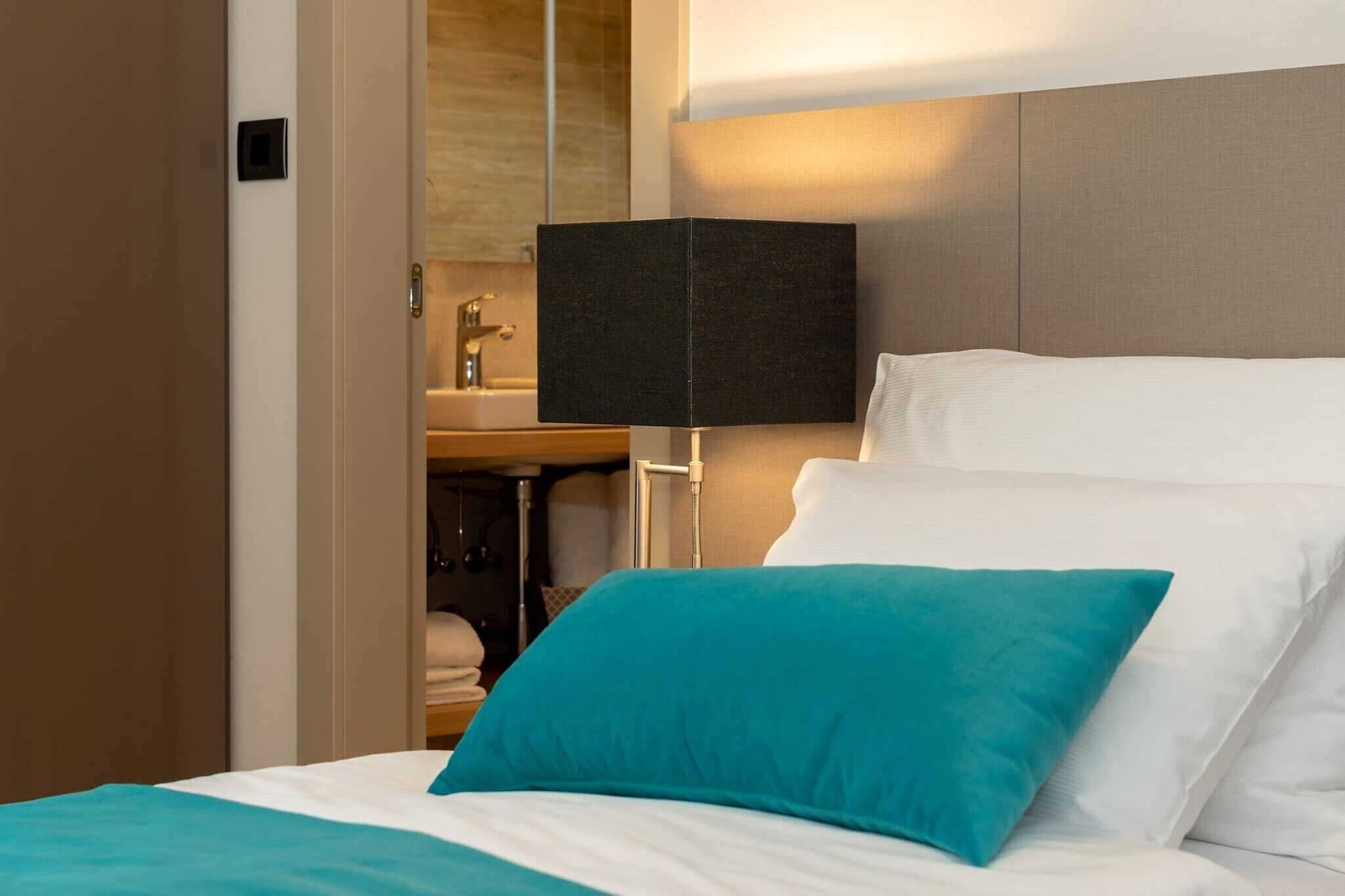 A neatly made bed with white and teal pillows, a black lamp, and a wooden nightstand with a mirror and a small sink.