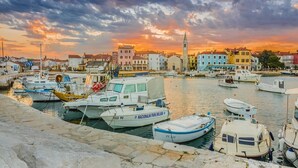A marina with various boats docked, a prominent building with a tower, and a sunset sky. - House in Fažana near Rock Beach (Fažana)