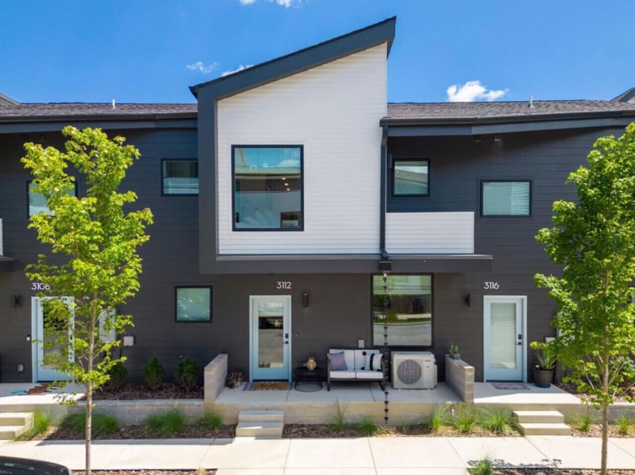 A modern two-story apartment building with numbered doors, a small outdoor seating area, and a well-maintained garden.