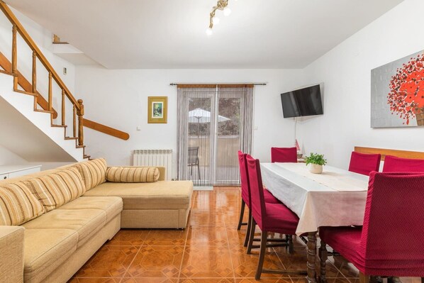 A living room with a sofa, a dining table with red chairs, a TV mounted on the wall, and a staircase.