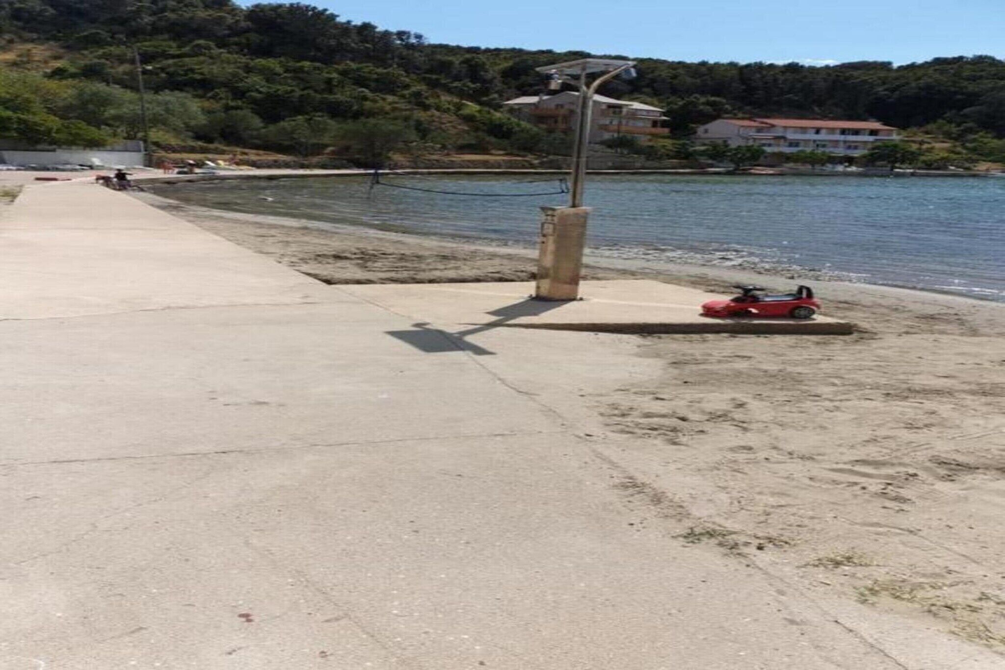 A concrete pathway beside a body of water, a red toy car, and a building in the background.