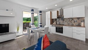 A modern kitchen with white cabinets, a built-in stove, and a tiled backsplash. There is a dining area with a white table and chairs, and a TV on a stand.