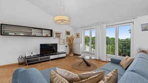 A modern living room with a flat-screen TV, a sofa, and a large window offering a view of greenery.