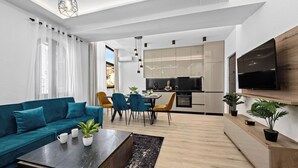 A modern living room with a teal sofa, a coffee table, and a dining area with chairs. A kitchen with a TV and cabinets is visible in the background. - Apartment in Pula near City Center Waterfront (Pula)