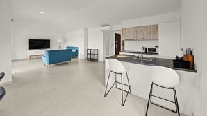 A modern kitchen with a central island, a dining area with a blue sofa, and a TV in the living room.