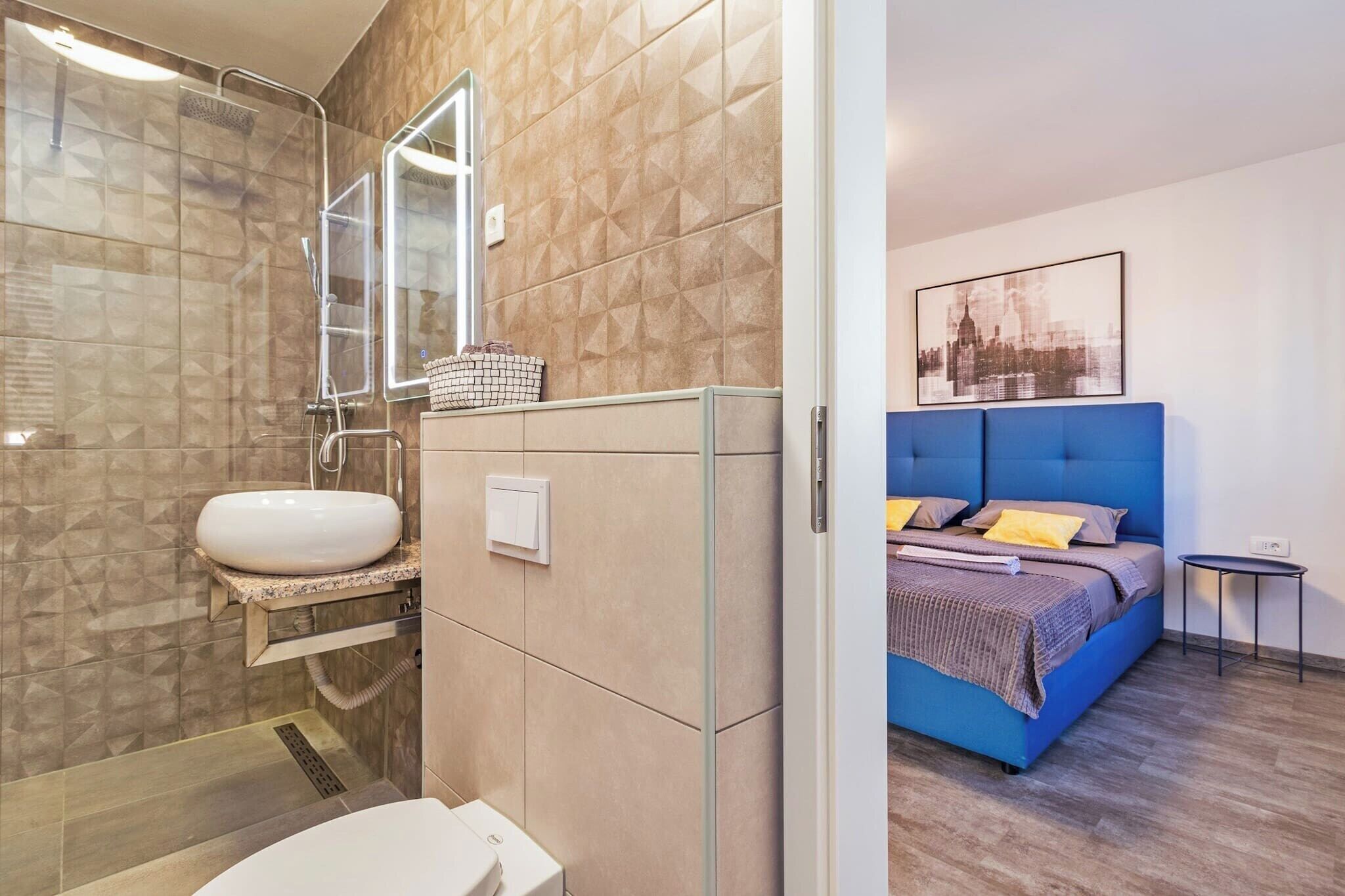 Ein modernes Badezimmer mit einer Duschkabine aus Glas, einem weißen Waschbecken und einer Toilette. Daneben befindet sich ein Schlafzimmer mit einem blauen Kopfteil, einem Bett mit Kissen und einem kleinen Nachttisch.