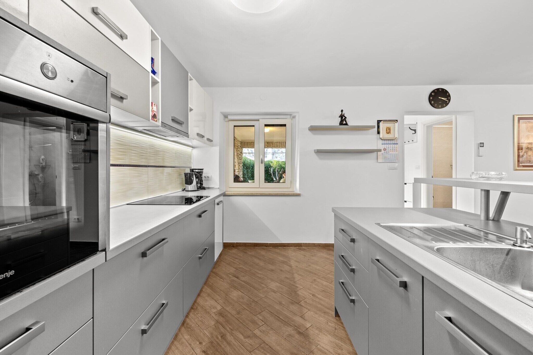 A modern kitchen with grey cabinetry, a built-in oven, and a window with a view of greenery.