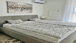 A modern bedroom with a large bed, a gray headboard, a wall-mounted air conditioner, and a window with sheer curtains. - Spacious apartment with wonderful sea view (Banjol)