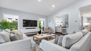 A spacious living room with a large flat-screen TV, a comfortable sofa set, a coffee table, and a view of the outdoors.