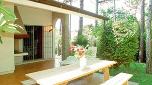 A wooden table with benches in a covered outdoor area with a view of trees and a building in the background. - Ground floor vacation home for six guests (Lignano Sabbiadoro)