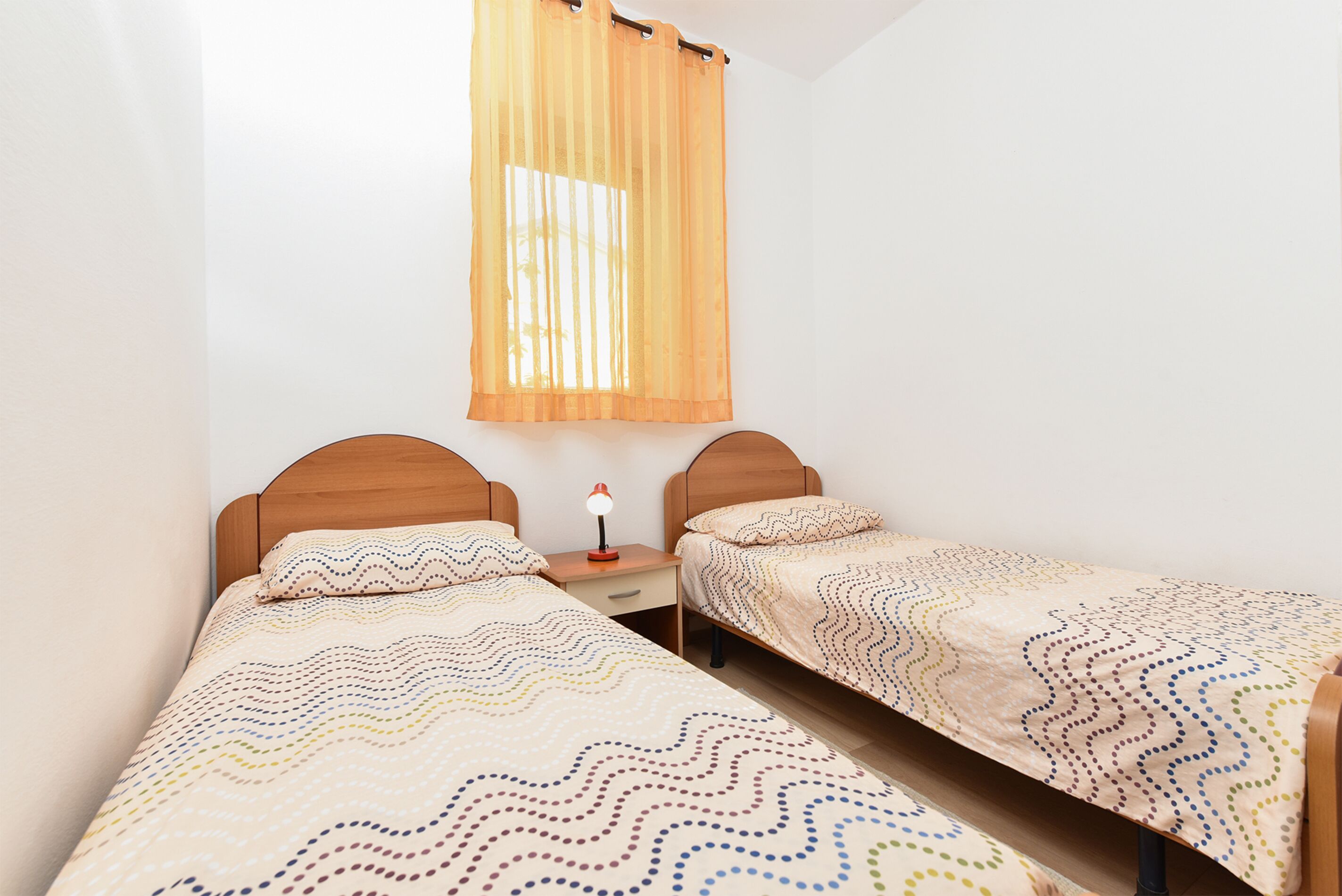 A room with two single beds, a nightstand, a lamp, and a window with sheer curtains.