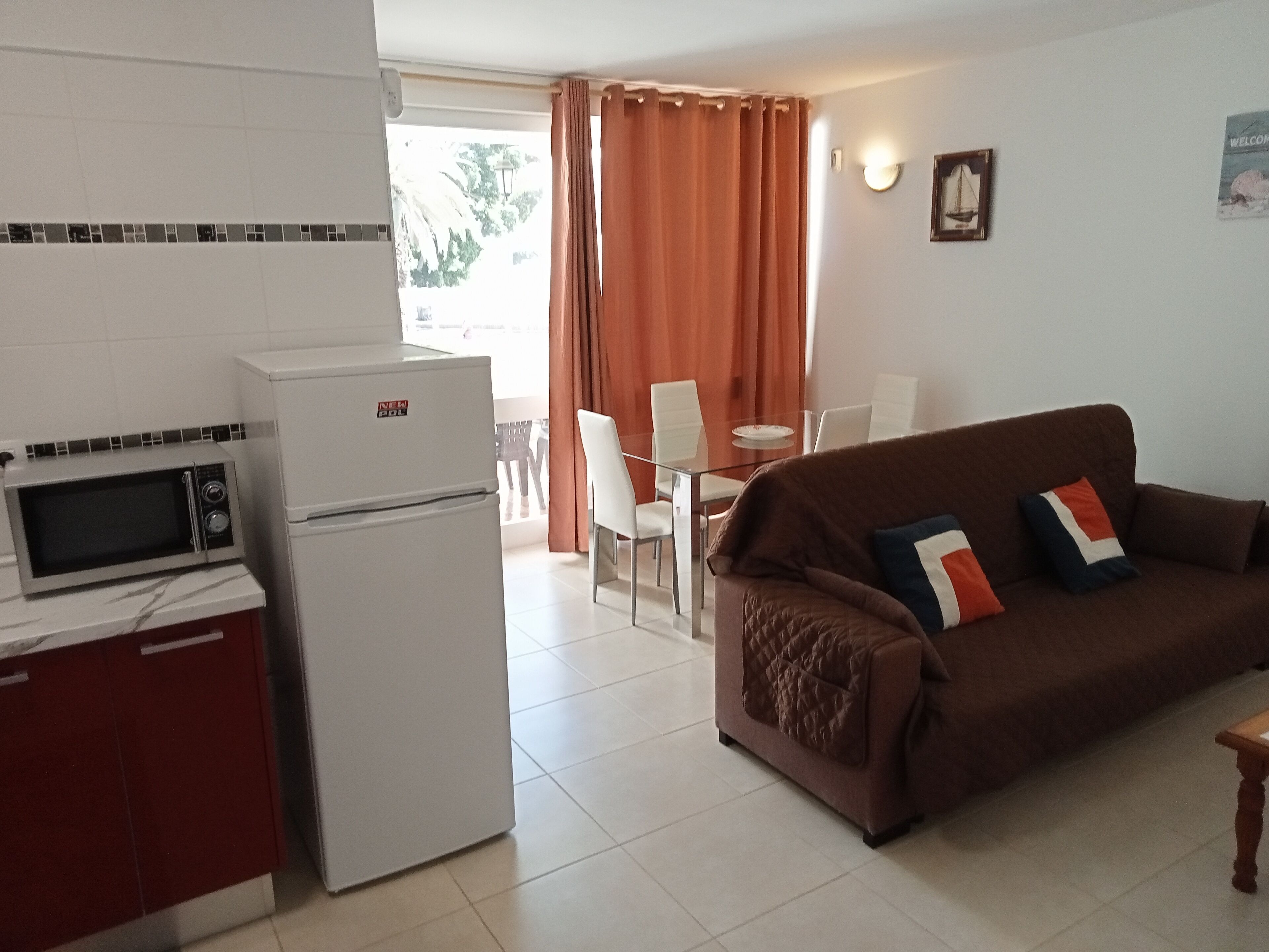 Un espacio habitable compacto con kitchenette, un sofá marrón con cojines, una mesa de comedor con sillas y una ventana con cortinas.