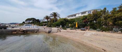 A coastal area with a clear water body, a sandy beach, and a building with a balcony overlooking the water.