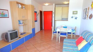 Un salon comprenant un téléviseur, un canapé, une table à manger avec des chaises, et un coin cuisine avec des placards et une porte rouge.