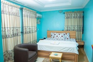 A bedroom with a bed, a chair, a bedside table, and curtains. - U.I HOTEL (Ibadan)