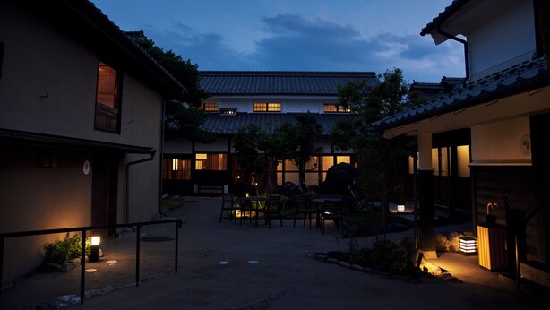 A traditional Japanese courtyard with a wooden table and chairs, surrounded by buildings with tiled roofs and illuminated windows. - Travel Stop UNNOWA (Tomi)