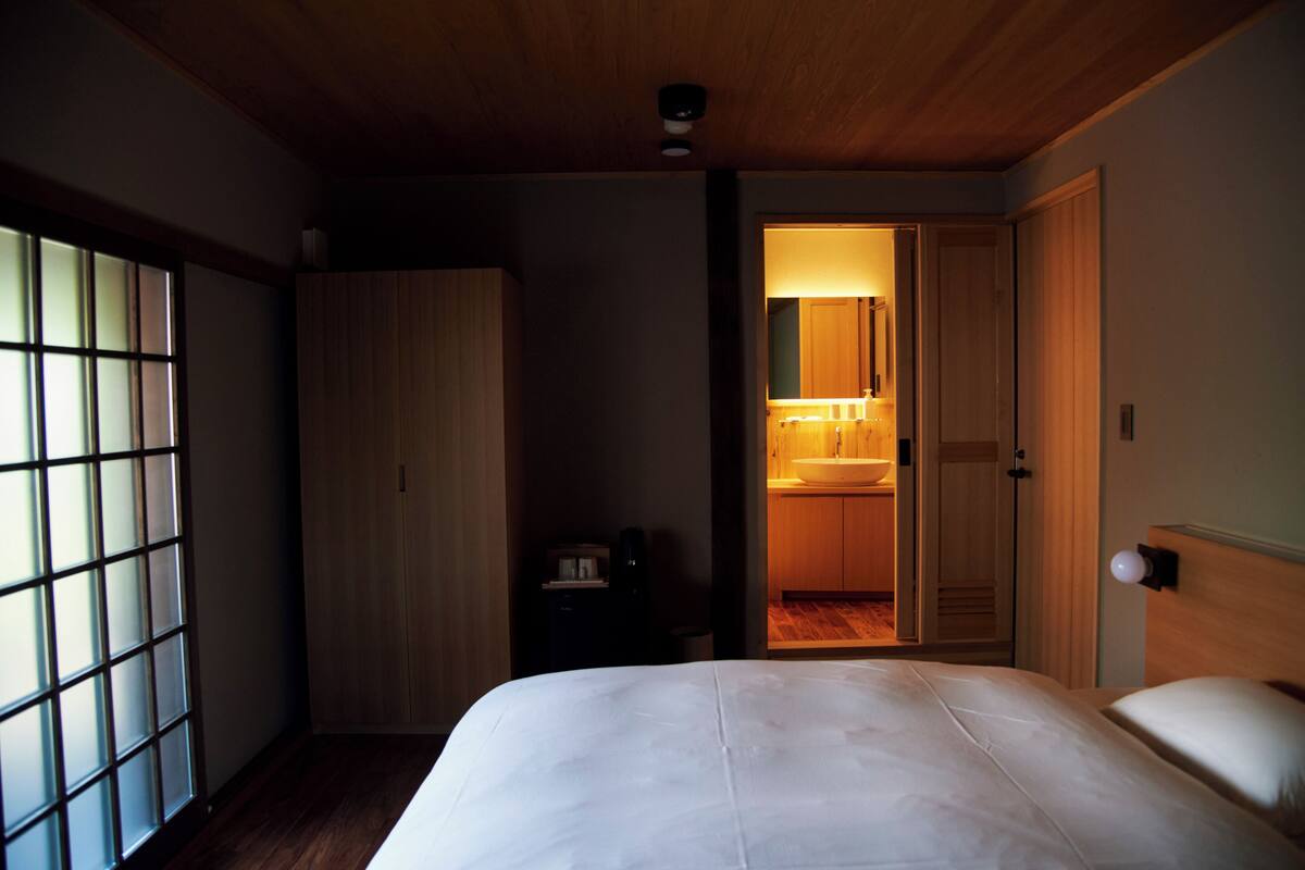 Hotellihuone, jossa on suuri sänky, vaatekaappi, pieni pöytä lampun kanssa ja näkymä kylpyhuoneeseen avonaisen oven kautta.