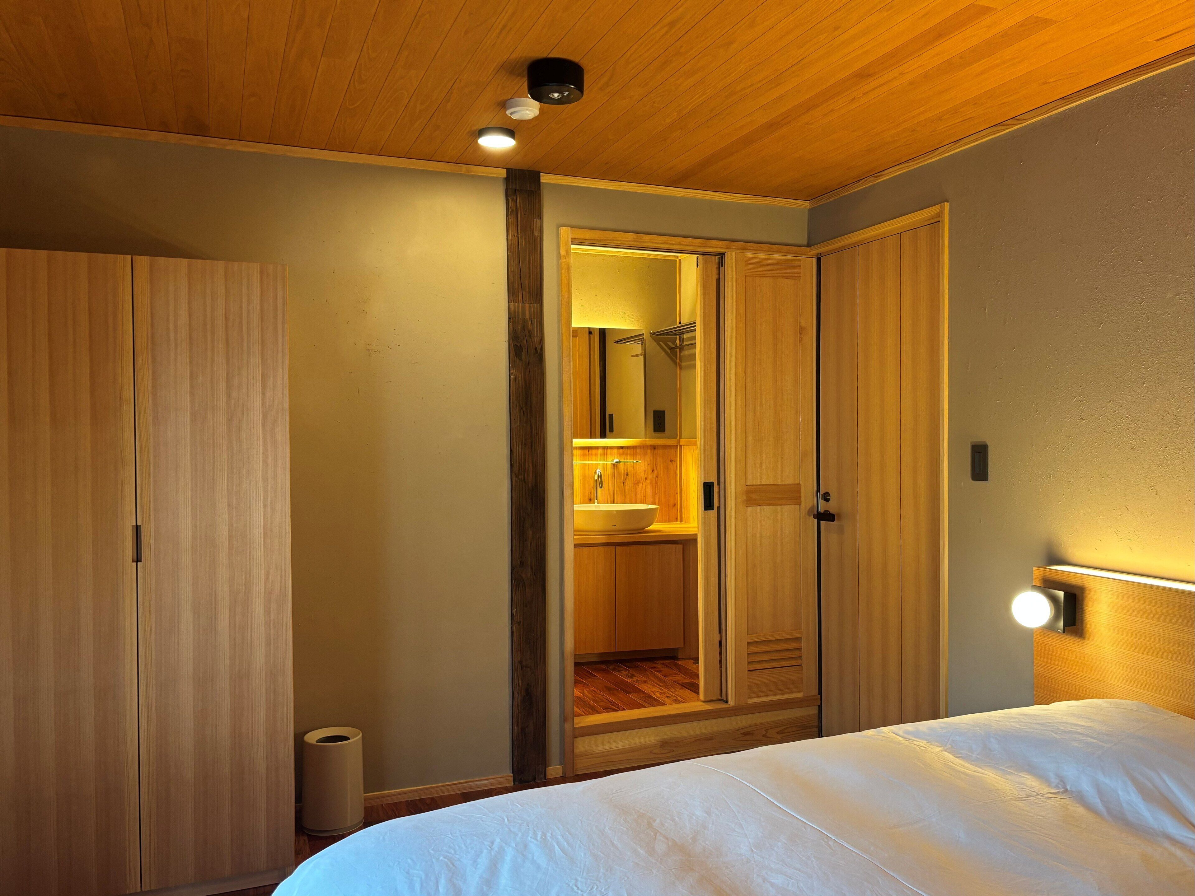 A bedroom with a wooden ceiling, a bed with white bedding, a wooden wardrobe, and a bathroom with a sink and mirror.