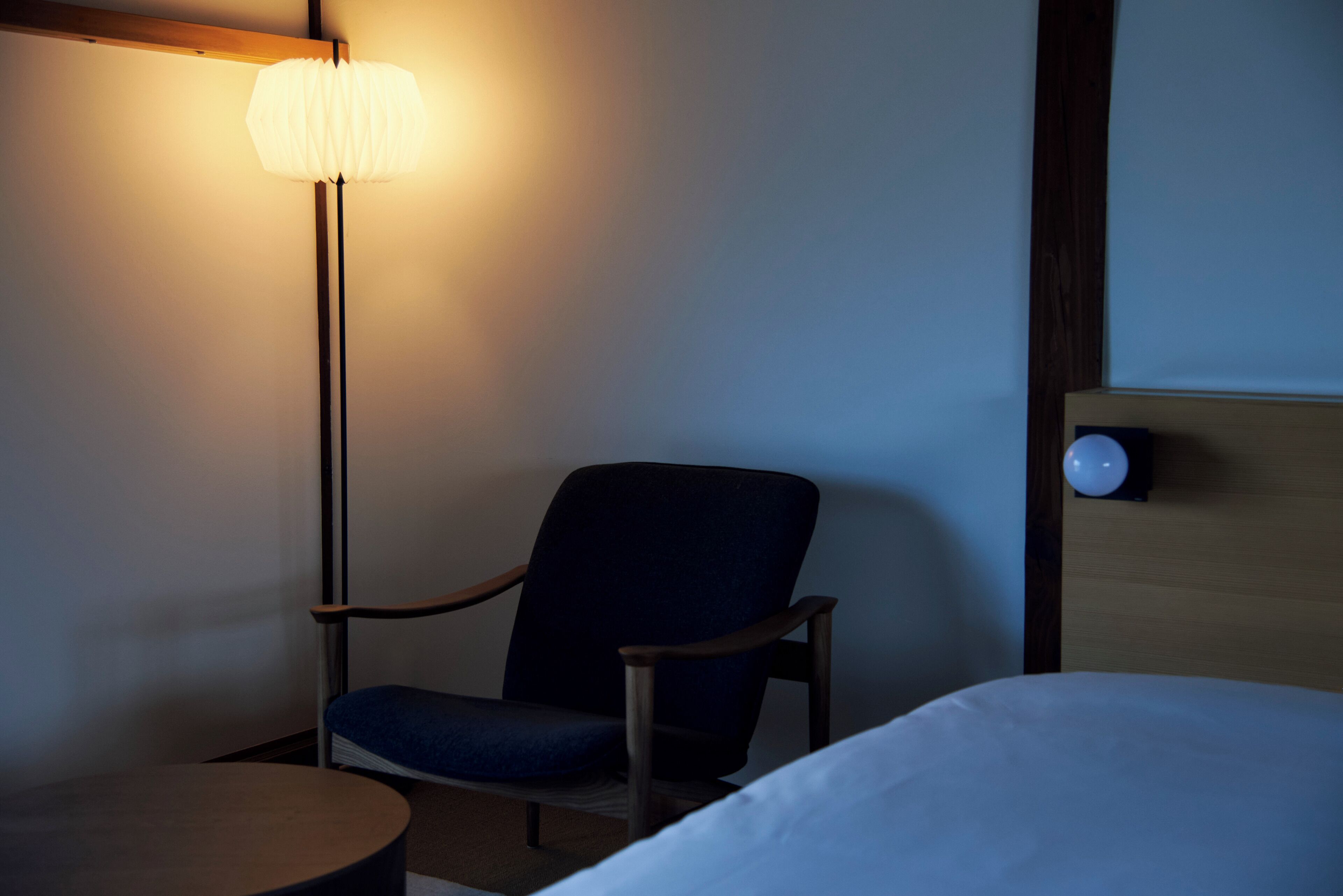 A hotel room with a single bed, a chair, a small table, and a floor lamp.