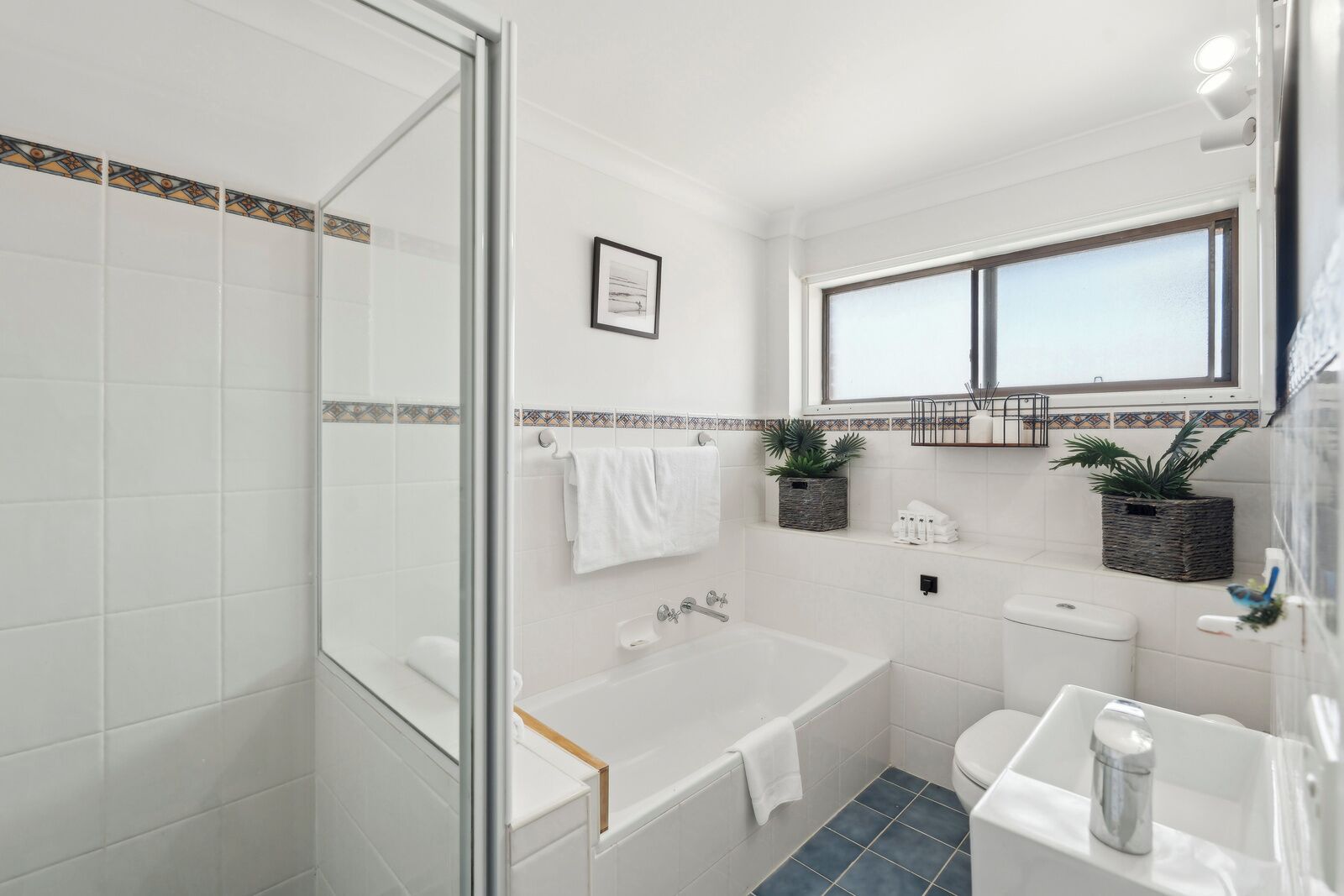 Une salle de bain moderne avec une douche à parois de verre, une baignoire, des toilettes et un lavabo. Deux plantes en pot sont posées sur le rebord de la fenêtre, et un tableau encadré orne le mur.