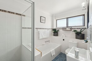 A modern bathroom with a glass shower enclosure, a bathtub, a toilet, and a sink. There are two potted plants on the windowsill, and a framed picture on the wall. - Barefoot on First - Affordable Family Apartment by the Beach (SAWTELL)